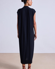 A woman stands against a neutral backdrop, wearing the "Olana Knit Midi" by Apiece Apart. The long, black sleeveless dress features a V-neck and is paired with elegant strappy red sandals, her hair styled in a graceful bun, creating an ensemble that radiates sophistication and extrafine Merino elegance.