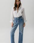 A person stands against a white background, effortlessly styled in Cissa's relaxed-fit Daily Shirt made from paper cotton with rolled-up sleeves, paired with comfy blue jeans. Their long brown hair complements the earthy tones of their brown loafers.