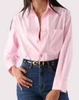 A person wears Cissa’s The Daily Shirt in light pink PAPER COTTON, tucked into high-waisted relaxed blue jeans with a black belt. With one hand on their hip, their face is partially out of the frame.