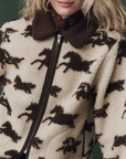 Someone wears The Pasture Jacket by The Great Inc.—a cream and brown relaxed-fit teddy jacket with a horse motif, brown collar, and visible zipper. The person’s face is partially out of frame.