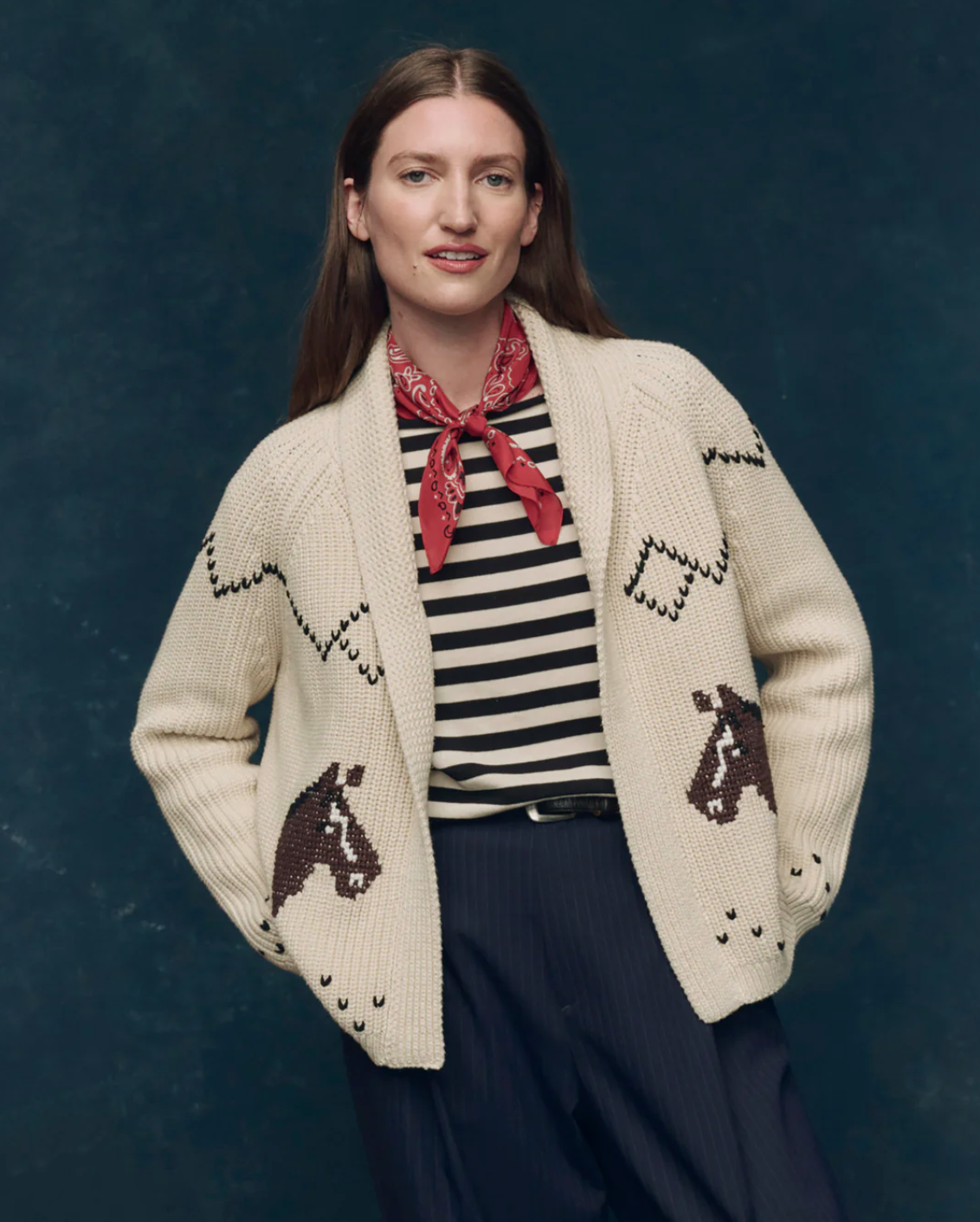 A person in The Great Inc.’s The Horse Lodge Cardigan—cream with hand-embroidered horses—paired with a black and white striped shirt, red bandana, and dark pinstriped pants smiles gently against a dark blue background, evoking western nostalgia.