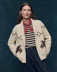 A person in The Great Inc.’s The Horse Lodge Cardigan—cream with hand-embroidered horses—paired with a black and white striped shirt, red bandana, and dark pinstriped pants smiles gently against a dark blue background, evoking western nostalgia.