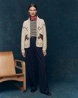 A woman stands before a dark blue backdrop, wearing The Great Inc.’s Horse Lodge Cardigan with hand-embroidered horses, a striped shirt and red scarf, and navy trousers. A wooden chair to her left adds subtle western nostalgia.
