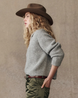 A person with long curly blond hair wearing The Great Inc.'s Crochet Collar Pullover, a brown wide-brimmed hat, and olive green pants with a brown belt, stands in profile against a neutral background.