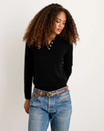 A woman with curly hair wears the Alex Mill Alice Polo Cashmere sweater in black and blue jeans, standing against a white background with her hands behind her back, looking down.