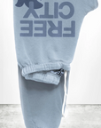 The FREECITY Large Sweatpant by Free City (sparrow, LLC) features a light blue color, dark blue "FREE CITY" print with bird graphic, unisex fit, white drawstring waistband, minimal design, and is proudly made in the USA.