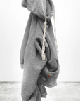 A gray BASIC GOODNESS HOODIE by Free City (sparrow, LLC)—a unisex light French terry pullover with white drawstrings—hangs on a hook against a white background and concrete floor. "freecity BASICgoodness" appears at the bottom.