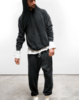 A man wears a loose-fit, made-in-USA Free City BASIC GOODNESS HOODIE with the hood up, matching dark sweatpants, white shirt sleeves, white socks, and black slippers, standing on concrete against a plain white background.