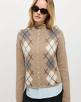 A woman in black sunglasses wears the Alex Mill Taylor Cardigan Argyle with pearl-like buttons over a light blue roll-neck sweater, standing against a plain background for a relaxed fit look.