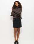 A woman with curly hair stands against a white background, wearing an oversized brown sweater layered over the Alex Mill Naomi Mini Skirt. She crosses her arms and gazes calmly at the camera.