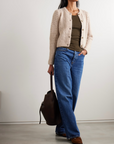 A woman wears FRAME's The Plush Cardi in beige over an olive top with blue wide-leg jeans, brown shoes, and holds a dark brown bag. She stands against a plain wall, her face partially out of frame.