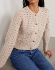 A woman wears FRAME's The Plush Cardi, a soft beige open-weave knit with gold buttons, paired with blue jeans as she sits on a light surface against a neutral background.