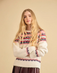 A woman with long blonde hair wears the Josie Sweater by Hunter Bell NYC, featuring colorful patterns, paired with a dark brown skirt, standing before a beige background.
