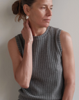 A woman with light skin and brown hair tied back wears The Sweater Rib Shell by Donni, a sleeveless ribbed gray top in a soft Tencel blend, as she touches her ear and looks thoughtfully to the side against a neutral background.