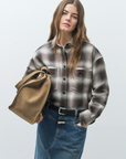 A woman with long, wavy brown hair wears the CLOSED Cropped Front OS Shirt and a denim skirt with a front slit, holding a large tan handbag. She stands against a plain light background, confidently facing the camera.