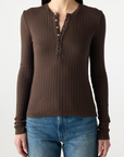 A woman wears the AMO Denim Lottie Top, a fitted ribbed henley in brown with metal buttons, paired with blue jeans against a light background. This cotton blend top is perfect for everyday style.