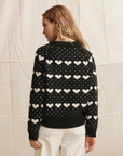 Someone with wavy, light brown hair models The Great Inc.'s The Heart Crew—a vintage-style black sweater with white hearts and dots—paired with light pants, against a neutral textured fabric backdrop.