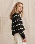 A young woman with wavy blonde hair wears The Heart Crew sweater by The Great Inc. and white pants, standing sideways and looking at the camera against a beige, textured fabric background.