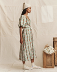 A woman stands sideways in The Fairground Dress by The Great Inc.—a floral-patterned white, green, and red midi with puffed sleeves—paired with a knit beanie and white high-tops, posing by wooden crates and flowers against a neutral fabric backdrop.