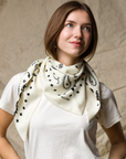 A woman wears the KUJTEN TIFA Scarf, featuring black patterns on cream, styled with a white t-shirt. She stands against a beige background, gazing softly to the side with a gentle smile.