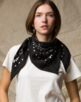 A woman with straight brown hair wears the KUJTEN TIFA Scarf, a black accessory with white patterns, draped around her neck over a white t-shirt, standing before a beige textured background.