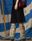 Wearing a Brazeau Tricot Crinkle Skirt, denim jacket, embroidered light blue cowboy boots, and holding a burgundy handbag, this person stands before a blue and white striped fabric backdrop.
