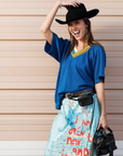 A smiling woman in the Aquarius Cocktail SIDE CAR mixed rib vee, a blue v-neck top with cropped sleeves, pairs it with a colorful skirt, black cowboy hat, black bag, belts, and jewelry as she poses against a beige wall.