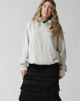 A woman with wavy blonde hair wears the Ping Pong Surf Club Le Surf Cowboy Hoodie in light gray, paired with a green scarf and a ruffled black skirt, standing against a plain light background.
