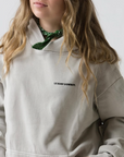 A person with long wavy hair wears the light-colored Le Surf Cowboy Hoodie by Ping Pong Surf Club, featuring "LE SURF COWBOY." on the chest and a green bandana. Their face is partially visible against a plain white background.