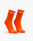 Two bright orange "Baby Steps" crew socks from Mother stand upright on a white background. The left sock features "MOTH" and the right sock "FUCK" in white text near the top.