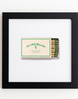 A framed box of matches with the label reading "The Old Warsaw, Dallas Texas 214-528-0032" hangs on a white wall. The matchsticks inside have green tips. The Art Square Blk Frame by Match South boasts a white flat profile and a white mat background, enhancing its minimalist, classic look.