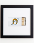 A **Match South Art Square Blk Frame** in a black square frame, featuring an acrylic design of a horseshoe and sword crossed, with the year "1887" beneath them. The matchbox is partially opened, revealing several wooden matches inside. The frame hangs against a white wall, making it a striking piece among other prints.