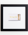 A minimalistic framed image with a white background displays the Art Square Blk Frame with partially visible matches inside. The matchbox, featuring gold cursive text that reads "The Beach Club Montauk," is positioned slightly off-center within the frame. Perfect for those who love elegant prints and sleek frames by Match South.