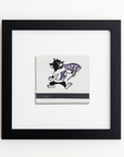 A black-framed picture, the Art Square Blk Frame by Match South, hangs on a white wall. The image inside the frame, an acrylic print, depicts a cartoonish wildcat holding a purple flag with the letters "K STATE" on it, representing Kansas State University.