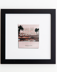 A framed picture displayed on a white wall shows a retro-style postcard with the text "PALM SPRINGS" in large lettering. The postcard, encased in an elegant Match South Art Square Blk Frame, features palm trees and a pool scene, giving off a vintage, summery vibe—perfect for fans of nostalgic prints.