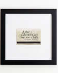 Framed artwork featuring a label from "The American Club Resort Hotel." The label is placed centrally and bordered by a black frame with a white mat. The design of the label includes elegant text and decorative elements, lending an air of sophistication to these Match South Art Square Blk Frame prints.