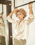 A woman with short blonde hair exudes modern elegance in the Celine Silk blouse by A Shirt Thing and olive pants, standing indoors holding a black bar above her head. She gazes confidently at the camera, her side reflected in a nearby mirror.