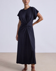 A woman stands against a plain background wearing the Apiece Apart Volta Twist Dress—an ankle-length, short-sleeved black organic cotton dress with a gathered twist waist—paired with brown strappy flat sandals.