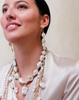 A woman in a cream silk blouse smiles gently, wearing long gold earrings and layered necklaces. Among them is the Bittersweet Designs Short Wrapped Pearls w/ Pearl Drop. She stands against a plain, light background.