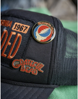 The MadeWorn Hat by Made Worn is a black mesh trucker cap with orange embroidery, an adjustable strap, and two limited edition pins: one with the Grateful Dead “Steal Your Face” skull and another with red/yellow “Grateful Dead” lettering.
