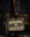 A person holds the Made Worn Artist Tote, a limited-edition olive-green cotton canvas bag with bold white "SPONSORED BY MADEWORN" text and colorful paint splatters; wooden chairs sit in the dim background.
