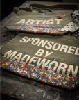 Two limited edition Artist Tote bags by Made Worn, crafted in olive-green cotton canvas with colorful paint splatters—one reads “ARTIST,” the other “SPONSORED BY MADEWORN”—are displayed on a paint-splattered surface.