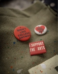 The Artist Tote by Made Worn features three custom buttons on cotton canvas: a red pin with "I DIDN'T LIKE COLLEGE ANYWAY," a small white heart pin, and a square red pin that reads "I SUPPORT THE ARTS.
