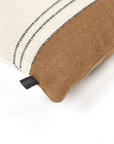 The Foundry Pillow by Libeco Home showcases a cream top with black wool-stitched lines and a solid brown bottom. A small black fabric tag accents the side of this rectangular artisanal pillow, which rests on a white surface.