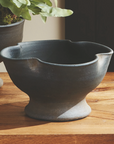The Kaloa Bowl by Napa Home & Garden, a dark ceramic planter with a flared base and matte finish, rests on a wooden surface. In the background, part of another handcrafted planter with green foliage is visible.