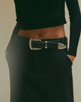 A person wearing a black ribbed long-sleeve top, black skirt or pants, and the B-Low The Belt Jordana Mini genuine leather Western belt with a large silver buckle stands with hands in their pockets.