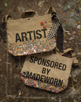Two beige cotton canvas Artist Tote bags by Made Worn sit on a paint-splattered surface. One reads "ARTIST" in colorful glitter, the other "SPONSORED BY MADEWORN," both sparkling with similar decorative accents.