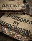 Two limited-edition beige cotton Artist Tote bags by Made Worn with colorful paint splatters—one features "ARTIST," the other "SPONSORED BY MADEWORN." The totes are displayed on a textured surface with custom buttons and a checkered cloth.
