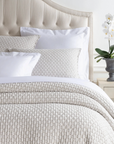 A neatly made bed features a beige Pinecone Hill Lodi Matelasse Coverlet, white pillows, and a tufted beige headboard. A potted white orchid rests on a light nightstand beside the bed against a white wall.