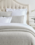 A neatly made bed features the Pinecone Hill Lodi Matelasse Coverlet and pillows in light gray, a tufted beige headboard, a white nightstand with a gray pot and white orchid, set against white paneled walls.